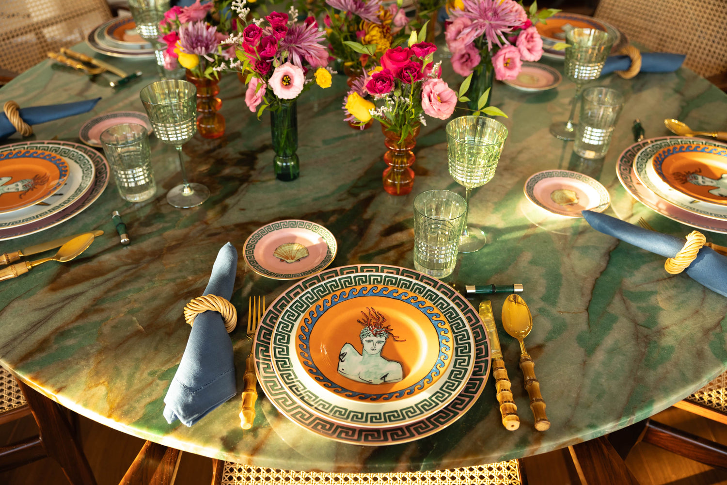 Mesa posta com pratos da coleção Viaggio di Netunno da marca Gionri 1735, taças e copos de cristal, talheres e guardanapos, sobre uma mesa de pedra com arranjos de flores coloridas no meio da mesa.