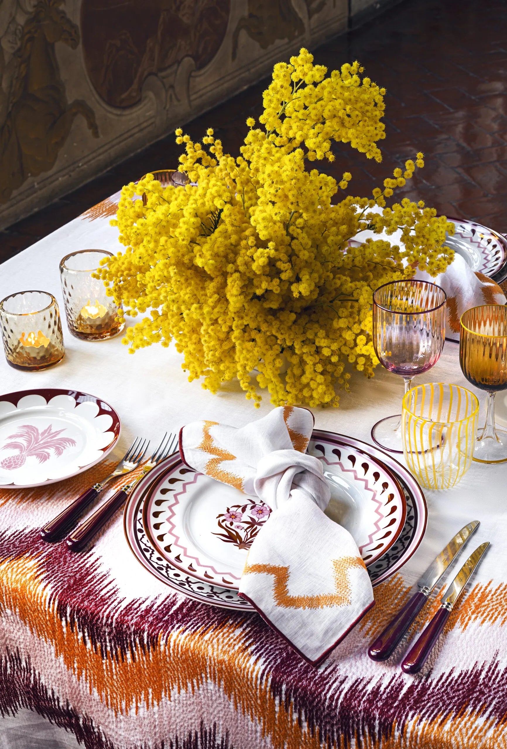 Mesa posta com pratos, taças e talheres, sobre toalha bordada e flores amarelas no meio.