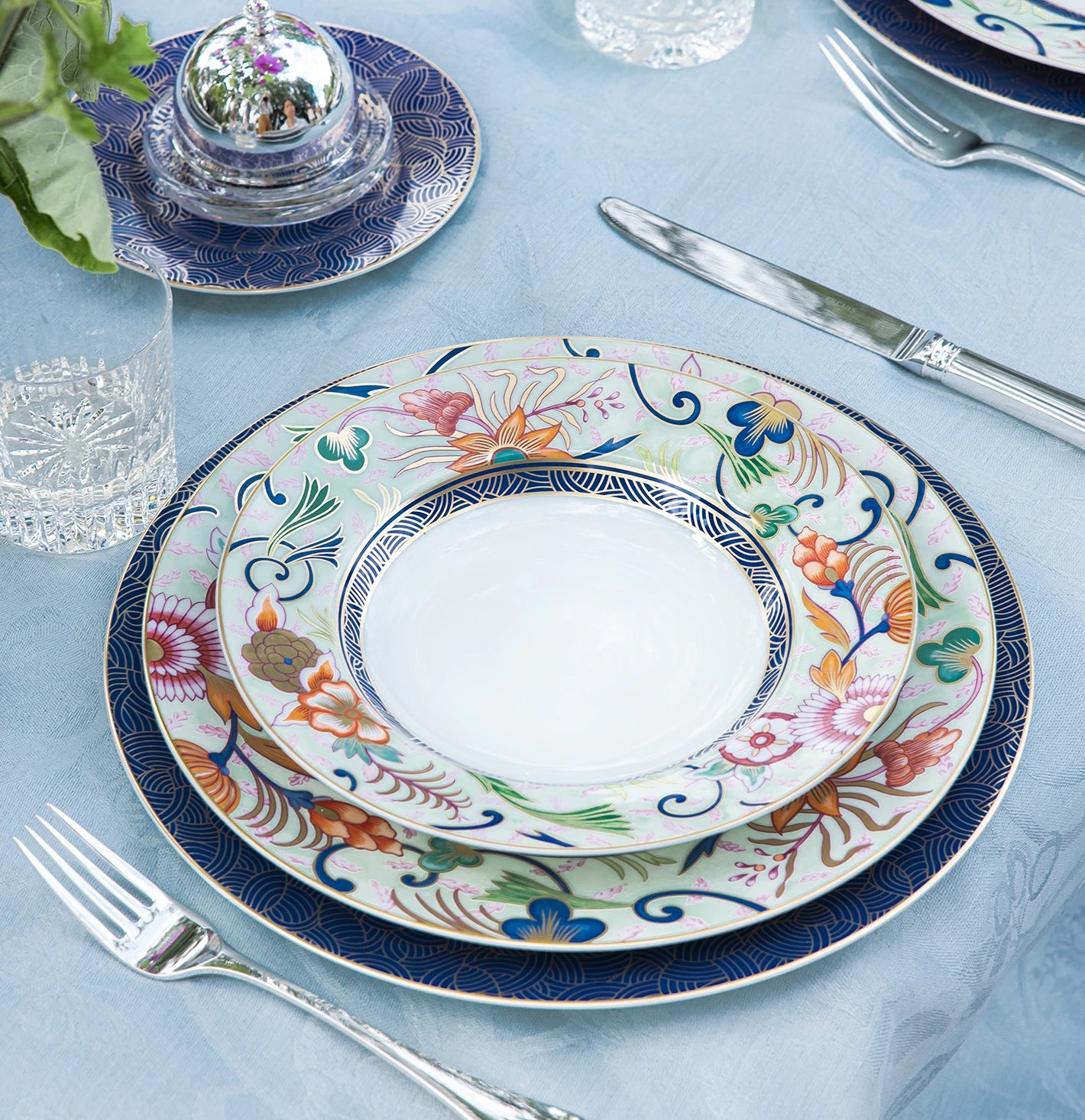 Mesa posta com pratos da coleção Imari, sobre toalha azul claro, com copos de cristal e talheres de prata.