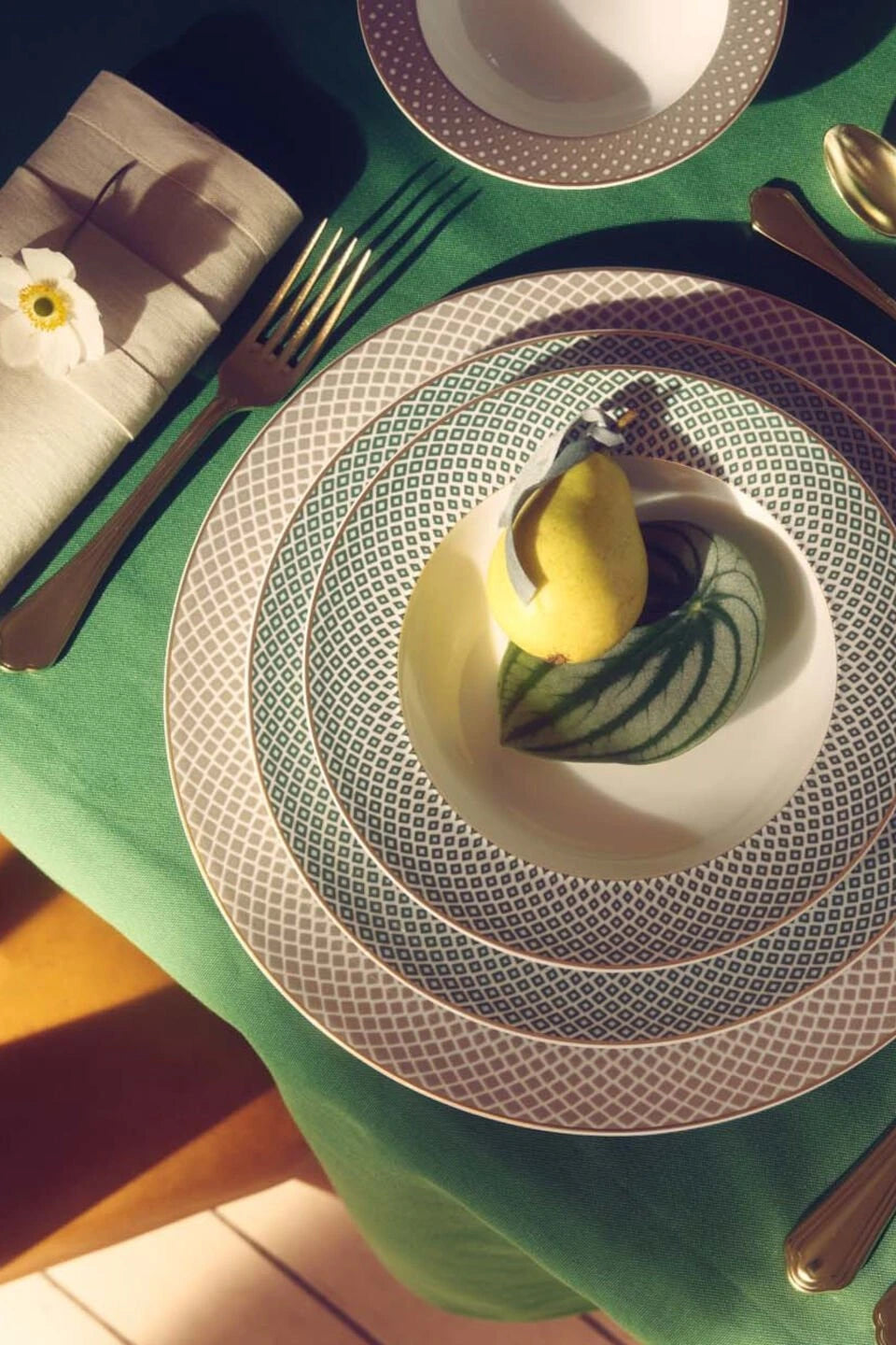 Mesa posta com pratos da marca Rosenthal, com talheres ao redor, guardanapo, sobre toalha de pano verde.