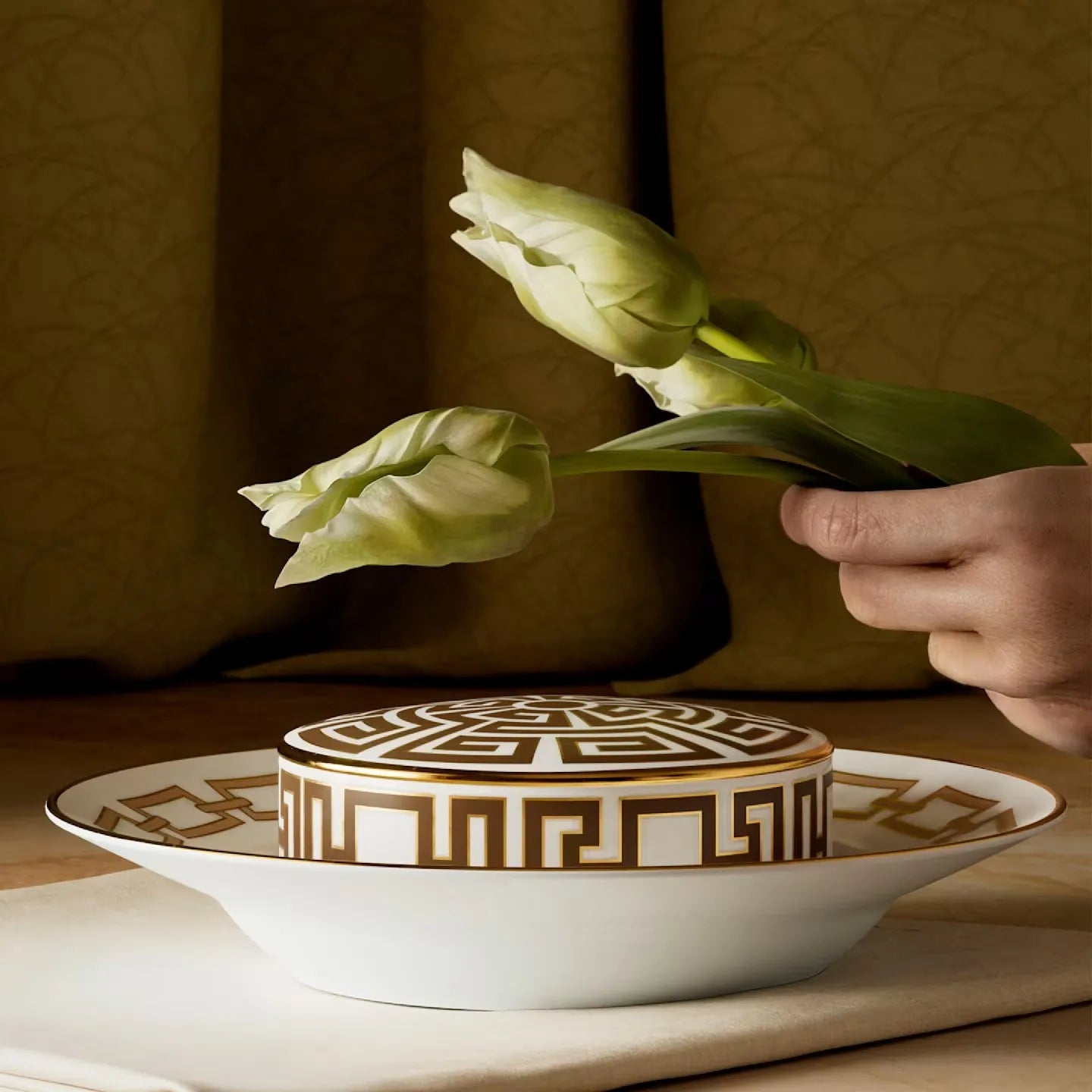Prato da coleção Catene e porta itens da coleção Labirinto, sobre uma mesa e uma mão segurando flores em cima.