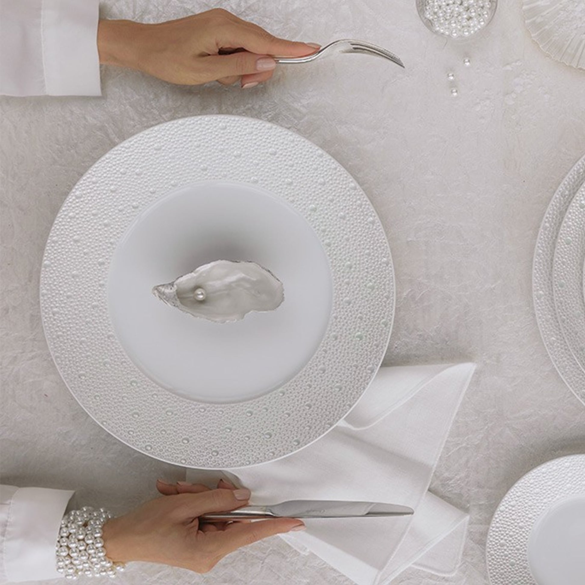 Uma mulher sentada à mesa, segurando talheres, com o prato Ecume em sua frente. Dentro do prato, está uma ostra com uma pérola dentro.