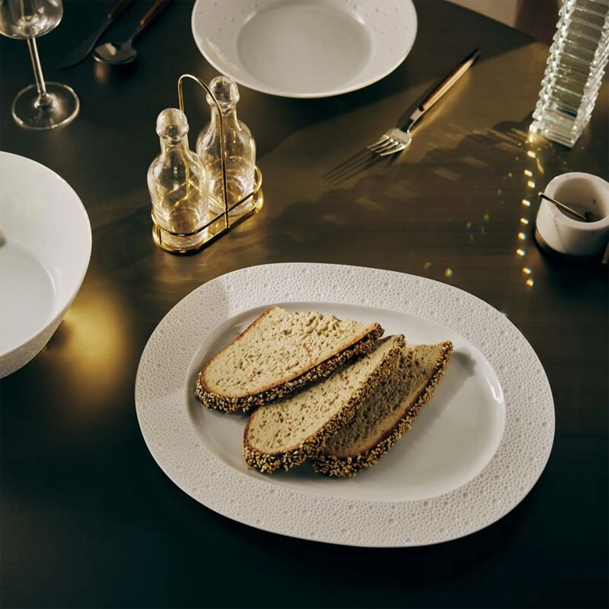 Travessa Ecume Branco 30 cm ambientada com pães, em cima de uma mesa preta, acompanhada por utensílios de mesa ao redor.