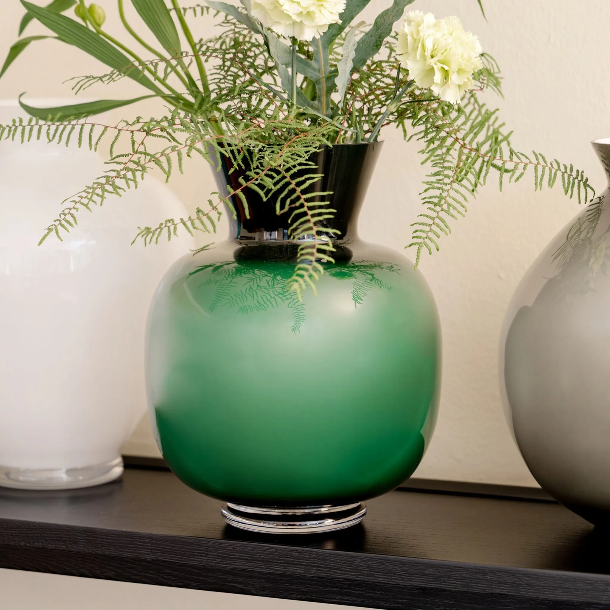 Vaso Rosenthal Anna Verde ambientado em prateleira de madeira preta, com flores dentro e ao lado de outros vasos.