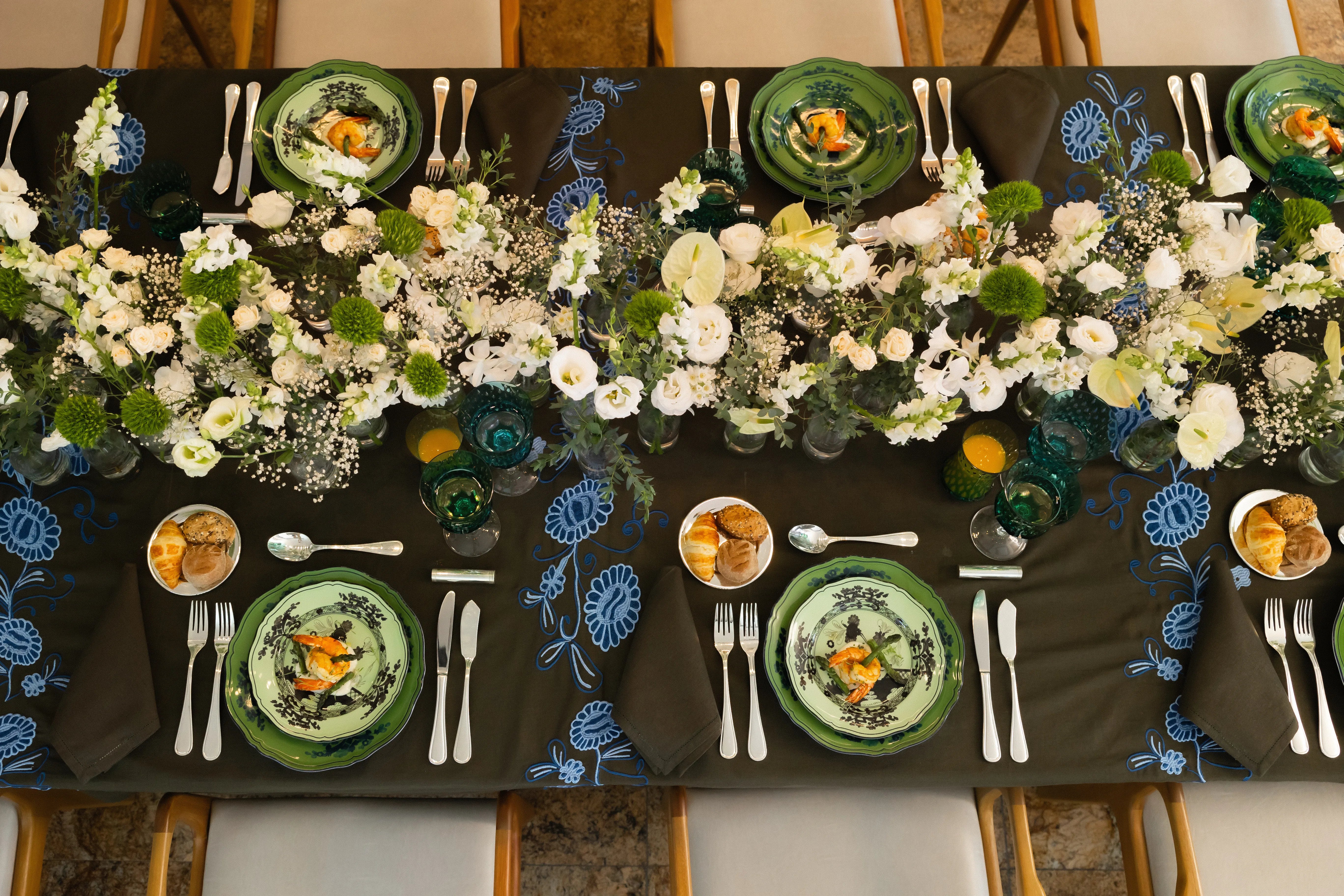 Mesa posta com pratos da coleção Oriente Italiano da marca Ginori 1735, sobre toalha bordada, com arranjos de flores brancas no meio da mesa.
