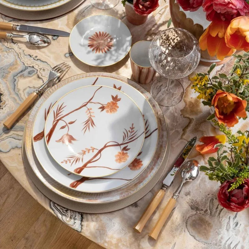 Mesa posta com pratos da marca Marie Dâage Paris, taça de cristal, talheres, sobre mesa de pedra, com flores laranja e rosa ao redor.