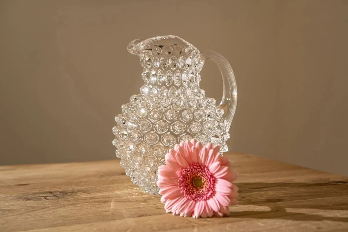 Jarra da marca Klimchi sobre mesa de madeira com uma flor rosa na frente.