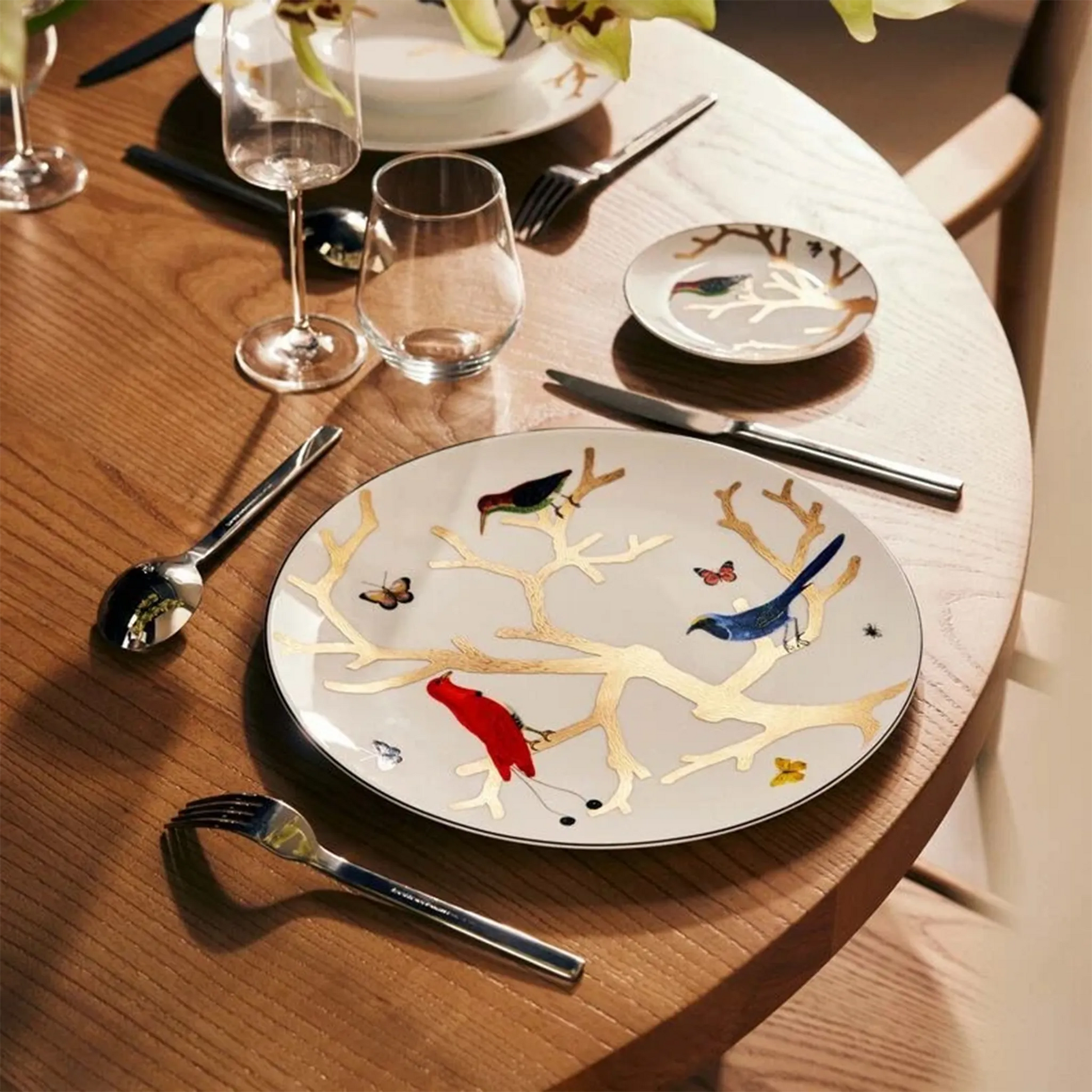 Sousplat Aux Oiseaux ambientado em mesa de madeira, acompanhado por talheres, taças e copo de vidro.