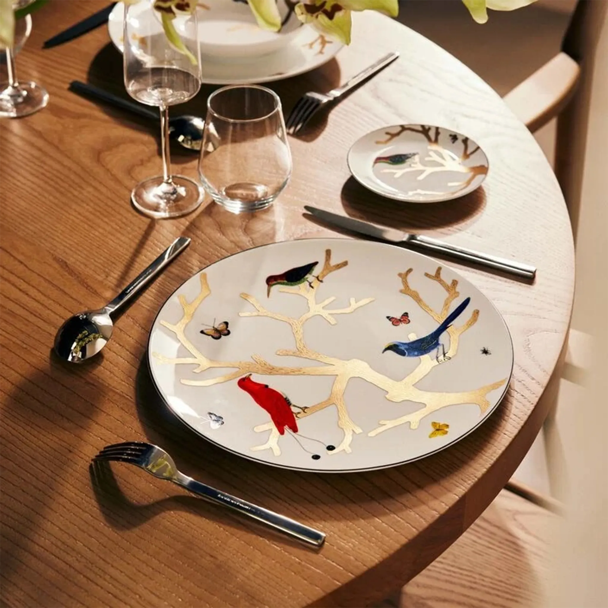 Sousplat Aux Oiseaux ambientado em mesa de madeira, acompanhado por talheres, taças e copo de vidro.