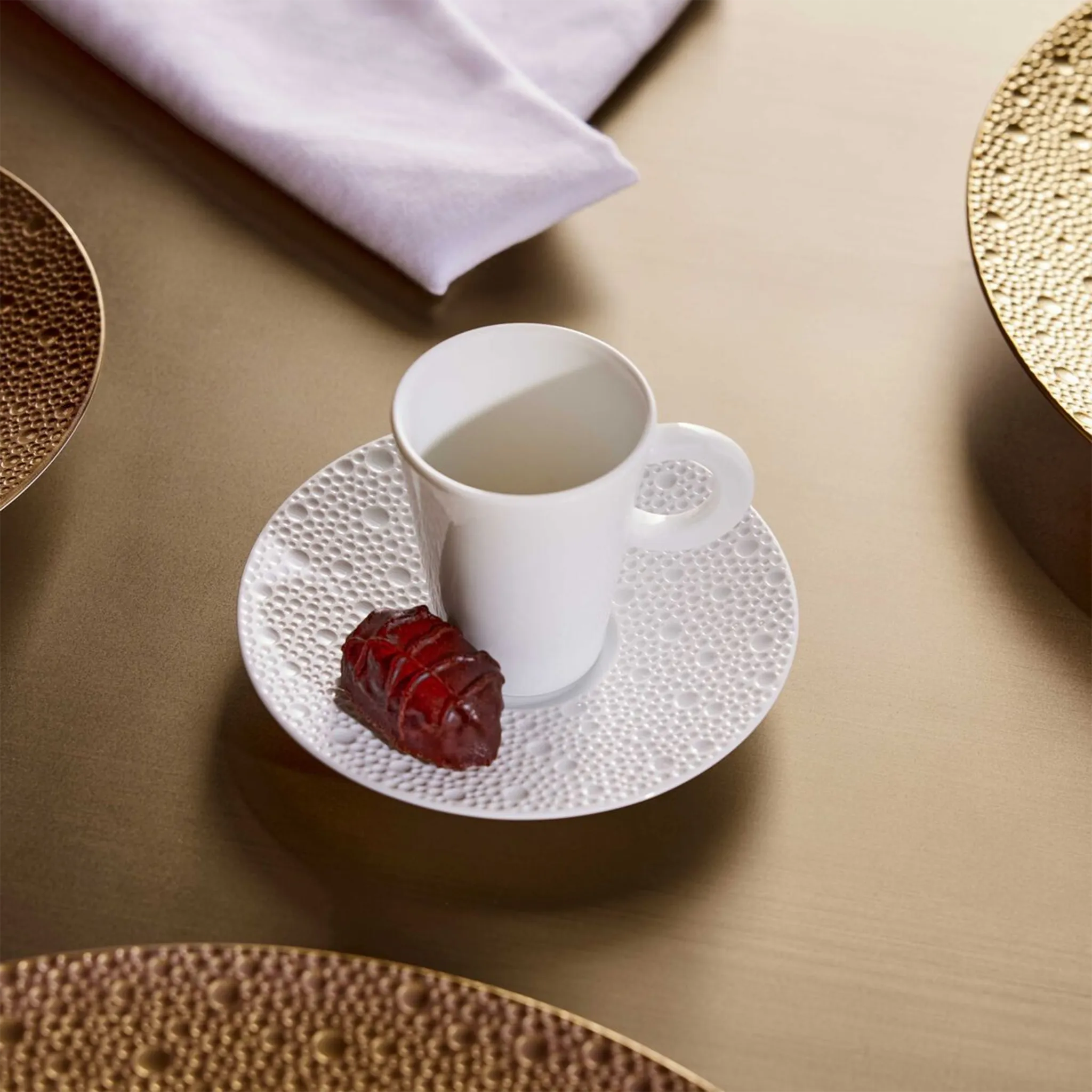 Xícara de Café Ecume Branco em cima de mesa, acompanhado com um pequeno doce e guardanapo.